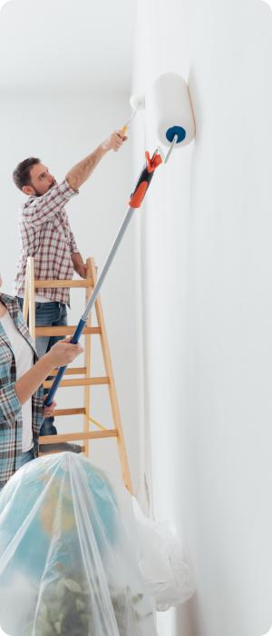 Two people painting a white wall, one using a roller and the other on a ladder.
